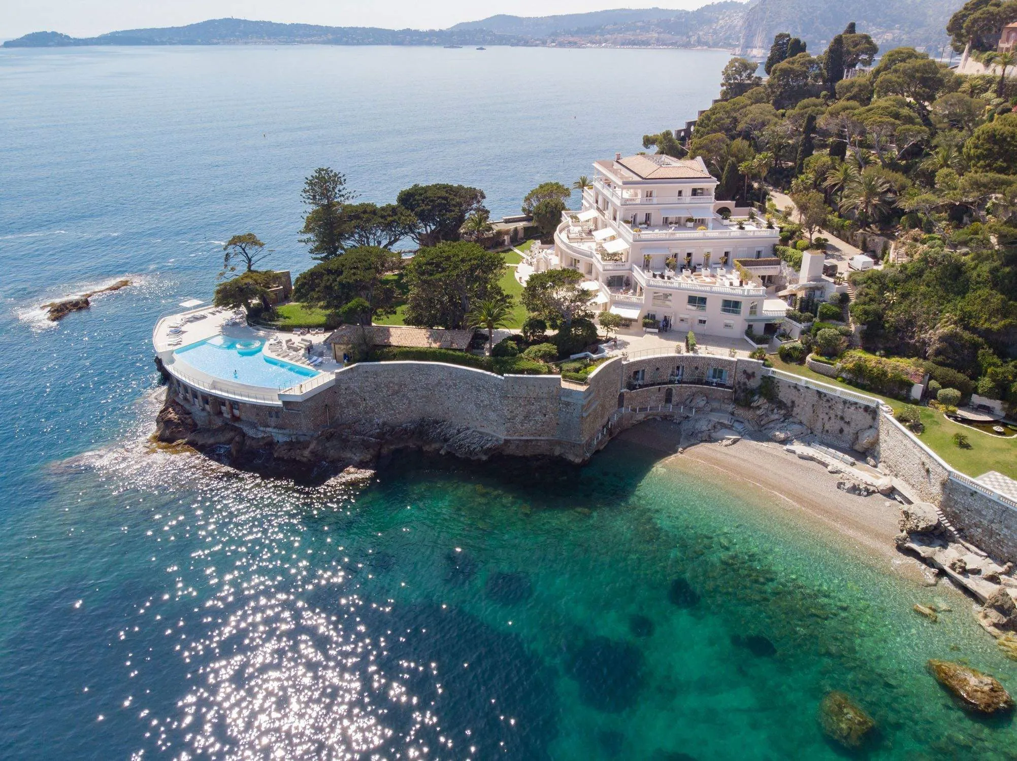 Cap Estel from the air, a white villa on its private peninsula above the seawater infinity pool and the pebble beach